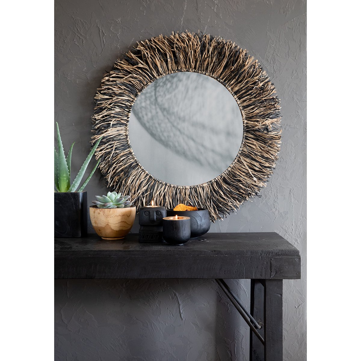 Round mirror with wicker frame on a dark wooden console table against a gray wall.